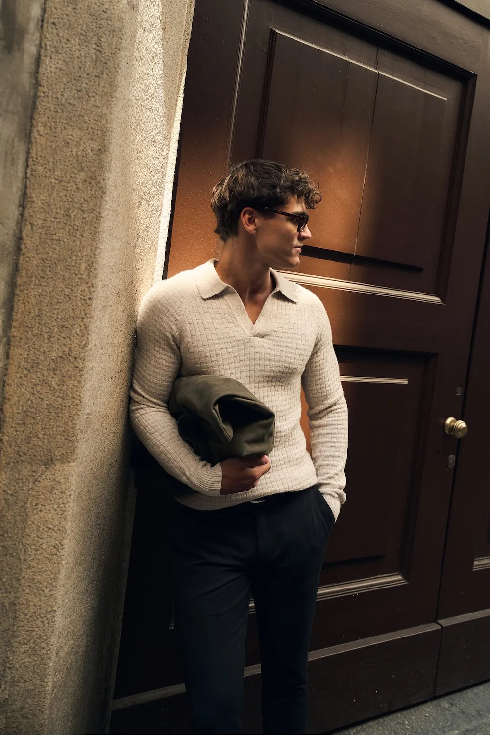 Man in a cream knit and dark trousers leaning in a stone doorway